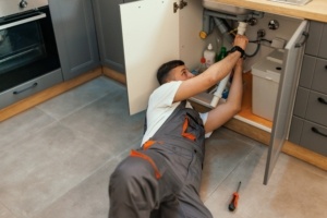 A young plumber is lying down and changing a pipe under a kitchen sink, with a screwdriver next to him.